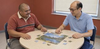 Two men in rust-red and light-blue shirts working on a landscape jigsaw puzzle at a beige table.