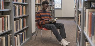 A young man in an orange-and-navy striped sweater reading a paperback on a red chair between library bookshelves.