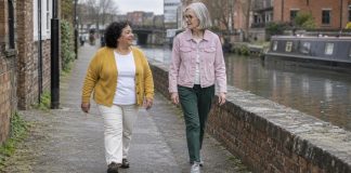 Two older women in mustard and pink jackets walking and talking beside a canal and a brick wall.