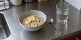 A bowl of oatmeal topped with banana slices, a spoon and a glass of water on a stainless-steel kitchen counter.