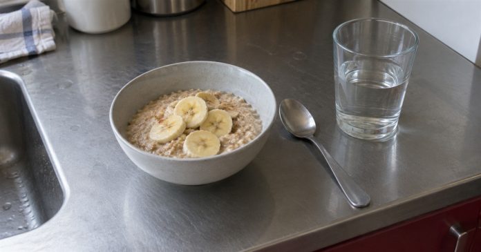 Self-love — everyday nourishment realistic cover 3496 A bowl of oatmeal topped with banana slices, a spoon and a glass of water on a stainless-steel kitchen counter.