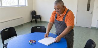 Man arranging blank drawing paper beside crayons on a blue activity table in a simple community room.
