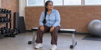 Woman in a pale-blue jacket taking a seated break on a padded bench in a brick-walled recreation room.