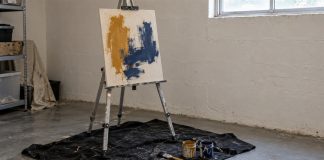 Unfinished ochre and blue painting on a metal easel with paint jars and a brush on a black drop cloth