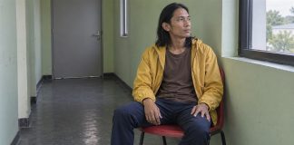 Man in a yellow jacket seated on a red chair beside a window in a quiet corridor