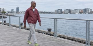 Man in a burgundy jacket and gray trousers walking along a waterfront boardwalk under cloudy skies