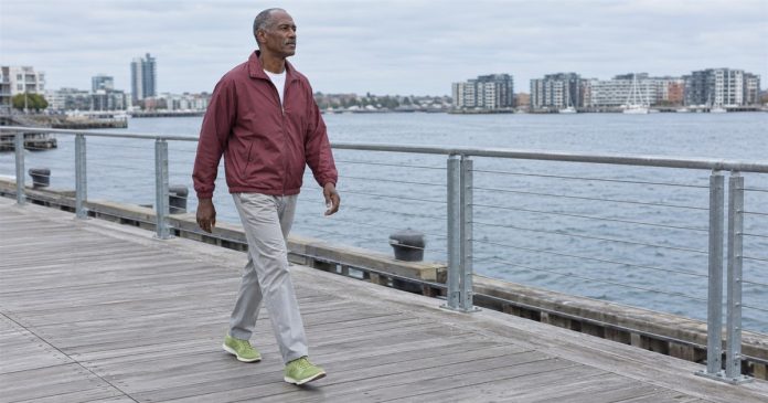 Man in a burgundy jacket and gray trousers walking along a waterfront boardwalk under cloudy skies