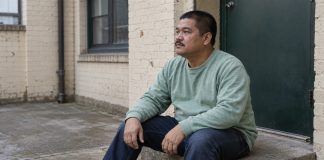 Man in a mint-green sweatshirt sitting quietly on a concrete doorstep in a brick courtyard