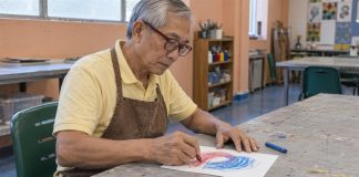 An older man wearing red glasses and a brown apron draws red and blue shapes on paper in an art room.