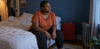 A woman in an orange shirt sits quietly on the edge of a bed, with a small closed phone on a red bedside cabinet.