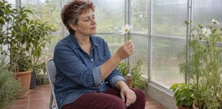 A woman in a denim shirt sits in a conservatory, looking at a white flower held gently by its stem.