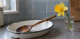 Worn wooden spoon in a chipped white enamel dish beside a yellow daffodil in a glass on a kitchen counter.