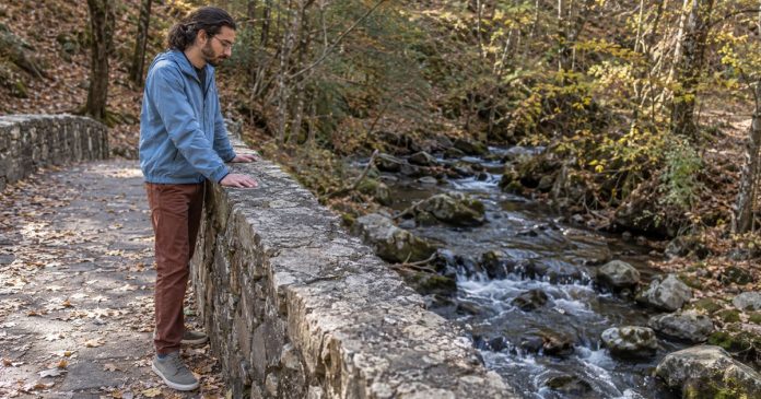 Spiritual growth — realistic creek reflection 3658 Man in a blue jacket resting his hands on a stone bridge parapet while looking at a woodland creek.