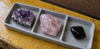 Amethyst, rough rose quartz and a polished black stone in separate gray tray compartments on a worn yellow workbench.