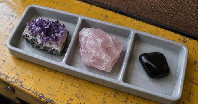 Crystal selection — realistic mineral tray 3670 Amethyst, rough rose quartz and a polished black stone in separate gray tray compartments on a worn yellow workbench.