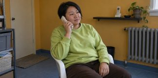 Woman in a lime-green sweatshirt listening on an ivory cordless phone in an ochre-walled room.