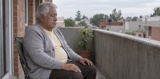 Man in a gray cardigan sitting with eyes closed and hands on his thighs on a concrete balcony.