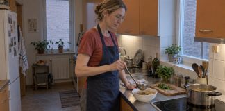 Woman in a blue apron serving soup into a white bowl in a modest orange-cabinet kitchen.