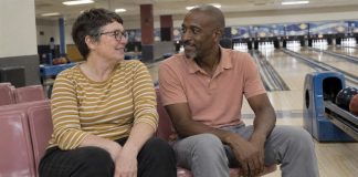 Two adults smiling at each other on pink seats beside bowling lanes.