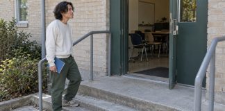 Man holding a blue exercise book on concrete steps outside an open classroom doorway.