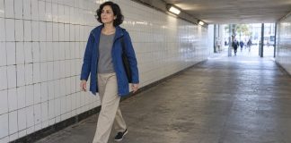 Woman in a blue raincoat walking through a tiled pedestrian underpass with daylight beyond.