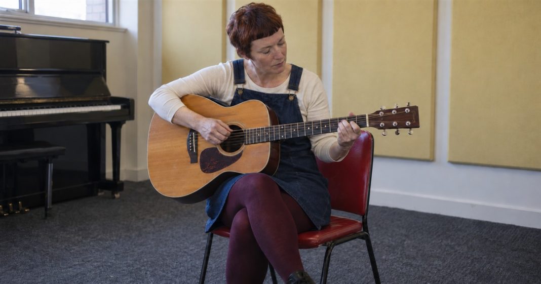 Woman in a denim dress practicing acoustic guitar on a red chair in a music room.