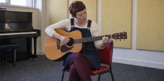 Woman in a denim dress practicing acoustic guitar on a red chair in a music room.