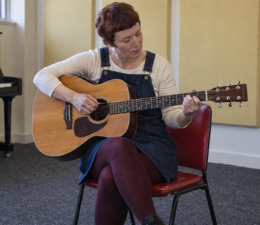 Woman in a denim dress practicing acoustic guitar on a red chair in a music room.