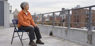 Older woman in an orange fleece sitting on a folding chair on an overcast urban rooftop.