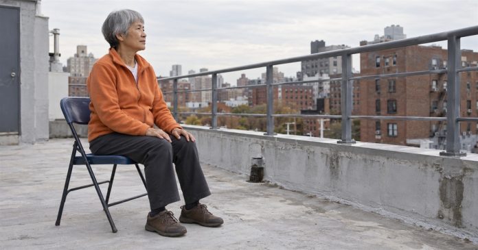 Old Energy realistic rooftop pause 20261004 Older woman in an orange fleece sitting on a folding chair on an overcast urban rooftop.