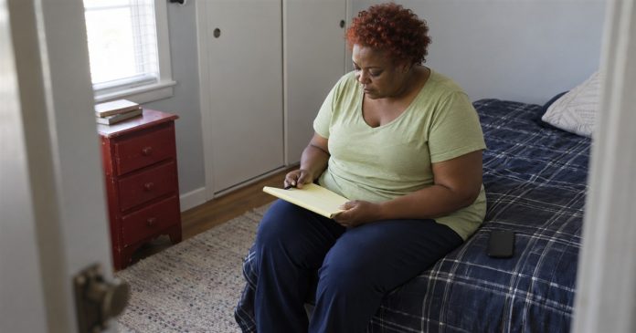 Woman in a pale-green shirt reviewing a yellow notepad on a bed, with a black phone beside her.