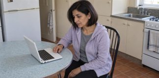 Woman in a pale-lilac hoodie preparing notes on a compact white laptop at a round kitchen table.