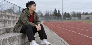 Woman in an olive jacket and red sweater sitting thoughtfully on concrete steps beside a running track.
