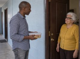 Man holding a closed meal container while speaking with an older woman at an apartment doorway.