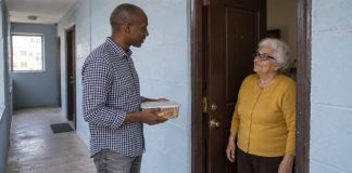 Man holding a closed meal container while speaking with an older woman at an apartment doorway.