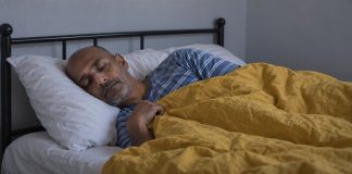 Man resting on his side with a white pillow and mustard-yellow duvet in a plain bedroom.