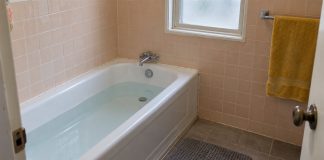 A plain-water bath in a peach-tiled bathroom with chrome taps, a yellow towel and a gray floor mat.