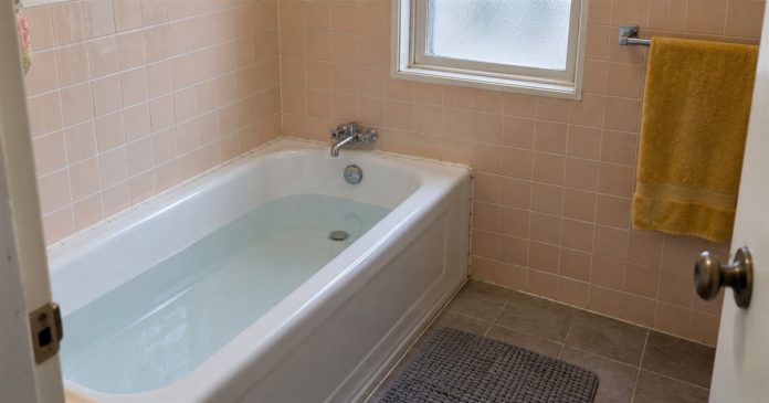 A plain-water bath in a peach-tiled bathroom with chrome taps, a yellow towel and a gray floor mat.