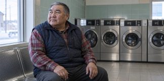 An older man in a navy vest taking a quiet pause on a metal bench beside a laundromat window.