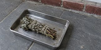 An unlit dried herb bundle resting in a metal tray on gray porch paving beside a brick threshold.