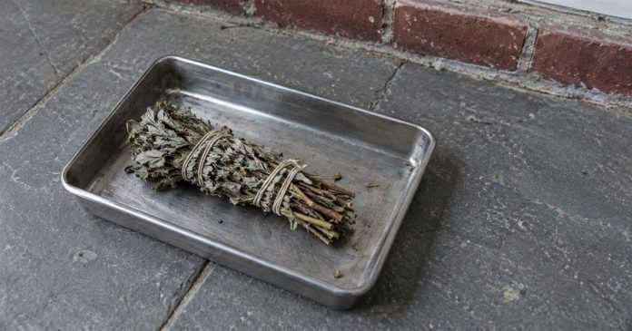 Psychic888 realistic unlit herbs porch 3873 An unlit dried herb bundle resting in a metal tray on gray porch paving beside a brick threshold.
