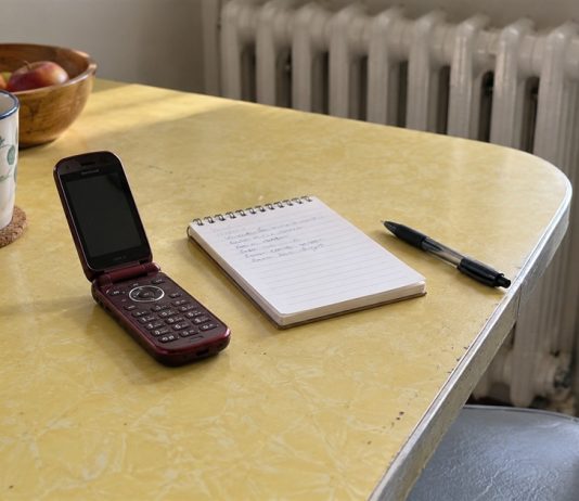 Burgundy flip phone beside a spiral notepad and black pen on a pale yellow kitchen table.