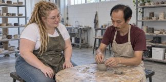 Woman watching a man shape a small clay pot at a worn round table in a ceramics workshop