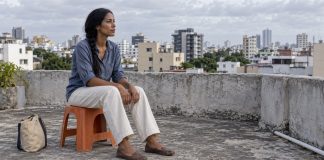 Woman in a blue shirt seated on an orange stool, looking across a city from a rooftop