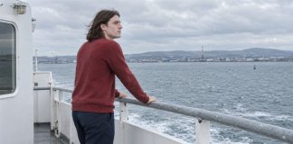 A young man in a red sweater looking across the water from a ferry deck