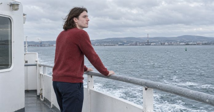 A young man in a red sweater looking across the water from a ferry deck