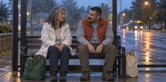 Two adults talking on a bus-stop bench at dusk with grocery bags beside them
