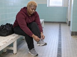 A woman tying a gray trainer on a worn white bench beside blue pool lockers