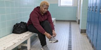 A woman tying a gray trainer on a worn white bench beside blue pool lockers