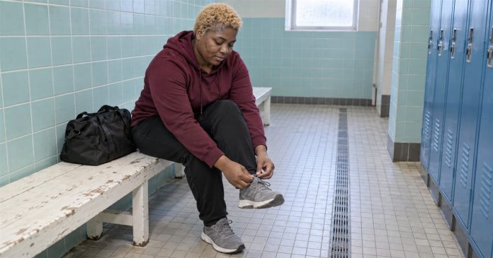 Realistic return to routine 3975 A woman tying a gray trainer on a worn white bench beside blue pool lockers